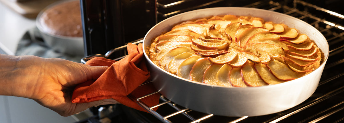 Una mano, che indossa un guanto da forno arancione, estrae dal forno una torta rotonda guarnita con fette di mela disposte con cura. Sullo sfondo, su una griglia, si intravede un altro dolce da forno.