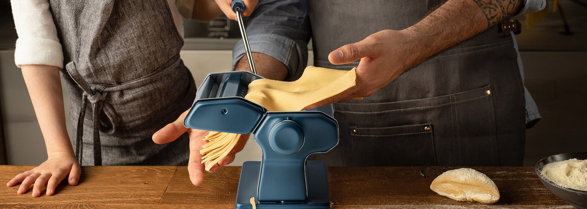 Due persone con indosso dei grembiuli utilizzano una macchina per la pasta per stendere e tagliare la pasta fresca su un tavolo di legno; nelle vicinanze si vede una piccola pila di impasto e farina.