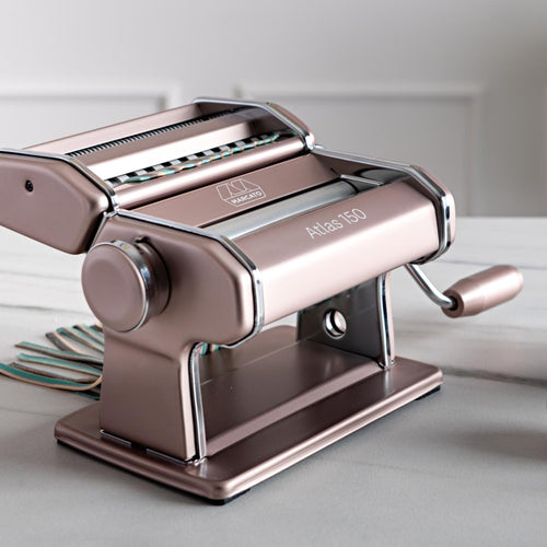 Una macchina per la pasta Atlas 150 color metallizzato è appoggiata su un piano di lavoro in marmo, con la manovella e i rulli ben visibili; è stata progettata per preparare la pasta fresca in casa.