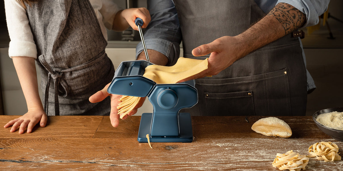 Due persone stanno usando una macchina per la pasta blu su un tavolo di legno, facendo passare l'impasto attraverso la macchina mentre ne escono delle strisce di pasta. Una persona gira la manovella, l'altra guida l'impasto. Sul tavolo ci sono farina e pezzi di impasto.